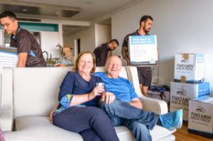 Happy older couple sitting on the couch while three men in Two Men and a Truck uniforms are moving and unpacking boxes.