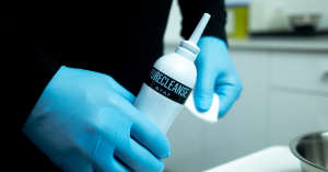 A clinician holding an opened bottle of Biomiq's PureCleanse STAT in one hand and gauze in the other. A stainless steel bowl shown on the table in front of the individual. Clinical setting.