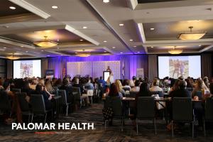 A conference room with attendees seated at tables, facing a speaker at a podium. Large screens display images and the Palomar Health logo is visible.