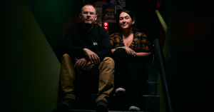 Robert Wood and Kyndra Vorster, the founders of Lucky 7, sit on a staircase in a dimly lit bar, exuding confidence and creativity. Kyndra wears a patterned top, while Robert sports a black sweatshirt and glasses, both showcasing their unique styles in the