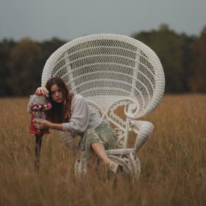 Woman seated on a large white wicker chair in a field, resting her head on a gumball machine.