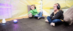 Two children wearing noise-canceling headphones relax on cushioned chairs in a sensory-friendly environment with soft lighting.