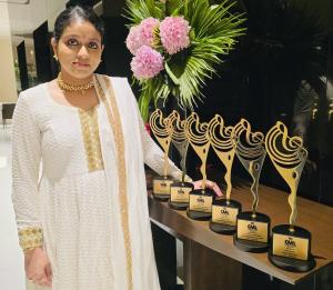 young lady is white traditional sari with 6 gold awards on shelf