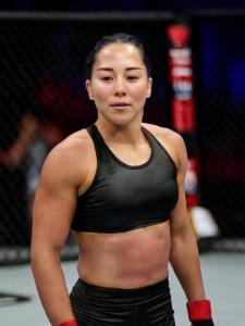 Sumiko Nakano, a determined female MMA fighter, stands in the octagon wearing a black sports top and shorts, showcasing her strength and readiness for competition.