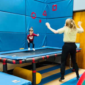 A therapist and a young child jump together on a trampoline in a padded therapy room.