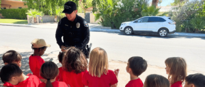 A group of children in red tops sitting and listening to a uniformed police officer outdoors.
