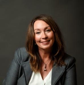 The photo is of Cynthia Adams (CEO and co-founder, Pearl Certification) with a warm smile, looking directly at the camera. She has long, light brown hair styled loosely around her shoulders. She is wearing a formal gray blazer over a white blouse and a si