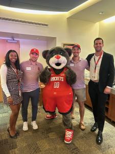 Business professionals from DOCUmation and The Houston Rockets pose together at an event, wearing red shirts and suits, alongside Clutch, the gray bear mascot.