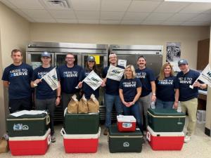 A group of DOCUmation employees wearing matching "Here to Serve" T-shirts pose for a photo. They are holding flags and standing behind coolers and bags of food, indicating they are participating in a community service event, donating food to Houston-area