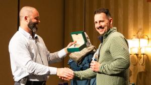 Hunter Woolfolk, CEO of DOCUmation, presents a Rolex watch to Chase Seamans, a consultant at DOCUmation Houston. Hunter is smiling as he hands over the boxed watch, while Chase, wearing a green jacket, smiles and shakes hands with him during the award pre