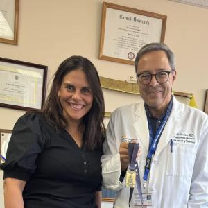Smiling woman with dark hair and eyes, wearing a black top, stands next to a smiling man with glasses wearing a white medical coat
