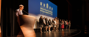 Person at podium with a group on stage under a projected event title at a pitch competition.