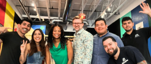 A diverse group of six individuals, smiling and posing for a photo in a brightly colored room with exposed ductwork and decorative walls.