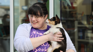 Christina Pascucci-Ciampa, a woman with dark hair and glasses, wears a purple shirt with the All She Wrote Books logo on it. She is holding her corgi, Ruby, as Ruby licks her face.