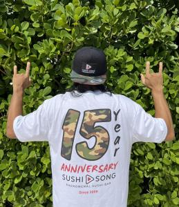 Chef Song wearing a "15 Year Anniversary Sushi Song" t-shirt with camouflage print, standing in front of green foliage. The shirt commemorates Sushi Song's 15 years in business since 2009, with the logo and tagline "Phenomenal Sushi Bar." The person is ma