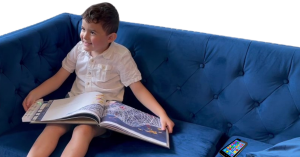 A child sits on a blue sofa reading a book and smiling while ignoring a smartphone on the sofa