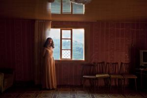 A teenage girl in a long, white formal dress stands alone by a window, looking into a sparsely furnished room. It is daylight outside and green hills can be seen outside the window,.