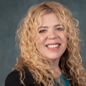 Portrait of DaVina Lara (CEO, bridgeMLS), a middle-aged Caucasian woman with curly blonde hair, smiling warmly. She is wearing a turquoise blouse and a black blazer. The background is a smooth, dark gradient.