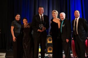 A group of six people dressed in formal attire pose together at an event. The man in the center is holding a clear award, smiling. They are standing in front of a dark curtain with blue lighting in the background, and musical instruments can be seen on a