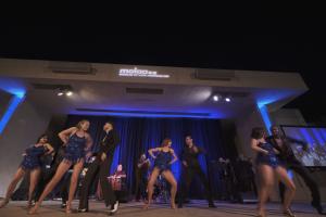 A group of dancers performing on an outdoor stage at night during a gala event. The dancers, wearing sparkly blue outfits and black suits, are mid-motion in pairs. The women are wearing fringe dresses, while the men are in sleek, formal dancewear. The sta