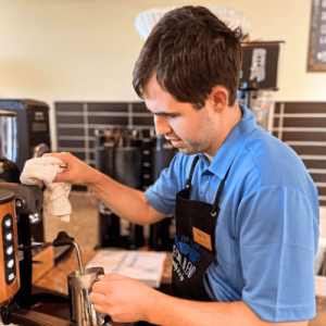 A barista wearing a blue shirt and apron carefully prepares a drink at a coffee machine.