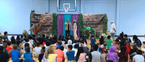 Children gather in a gymnasium to watch a live dinosaur-themed performance with actors and lifelike dinosaur props on stage.