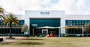 Exterior view of the link building in Ponte Vedra, Florida, a modern community hub for learning, coworking, and events. The building features a sleek white facade with large glass windows, shaded seating areas, and colorful outdoor furniture. Surrounded b