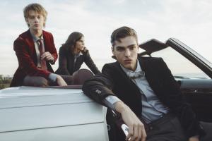 Three people in formal attire pose around a white convertible car against a cloudy sky background.