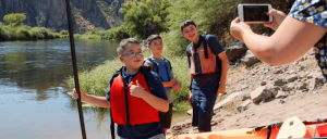 Three boys wearing life vests pose by a river while a person takes their photo, ready for a kayaking adventure in a scenic, rocky landscape.