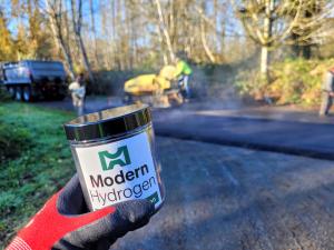 A close-up of a gloved hand holding a jar labeled "Modern Hydrogen" in the foreground, with a road paving scene in the background. Workers and machinery are applying asphalt on a rural road surrounded by trees and greenery.