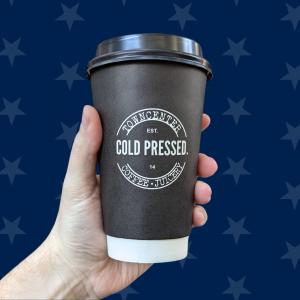 A hand is shown holding a black cup of locally roasted coffee from Town Center Cold Pressed in front of a dark blue background with faded stars.