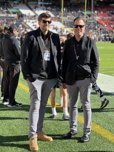 Two male physicians standing on the sideline of the Cleveland Browns football game wearing new medical kits that include grey pants and a black blazer.