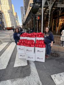 Two Operation Gratitude team members stand beside stacked boxes filled with red care packages on a New York City street, with buildings and holiday decorations in the background.