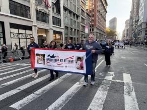 Participants march in the Veterans Day Parade, holding a banner that reads “Operation Gratitude – Saying ‘Thank You’ to All Who Served," with bystanders and buildings in the background.