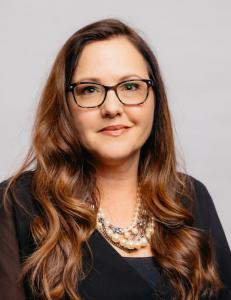 Portrait of Trish Martin, a Caucasian woman with long, wavy brown hair, wearing black rimmed glasses and a multi-strand pearl necklace. She is dressed in a black blouse and has a thoughtful, professional expression.