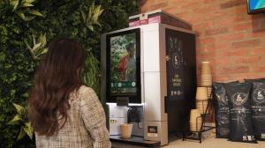 Employee waiting at an &BACK COFFEE bean-to-cup machine, waiting for a fresh cup of coffee to brew while watching an impact video playing on the coffee machine screen.