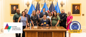 A group of people, including government officials and representatives, stand behind a seated official holding a certificate, with flags in the background and logos for the Autism Foundation of Oklahoma and IBCCES Certified Autism Center™ visible in the fo