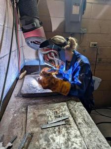 Sea Cadet wearing protective welding gear, including gloves and a helmet with face shield, concentrating on welding training at a workbench in a well-ventilated workshop.