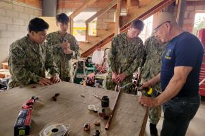 Sea Cadets in uniform participating in SeaBee training, gathered around a workbench while an instructor demonstrates proper soldering techniques with copper pipes in a workshop setting.