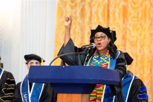 DBA graduate delivering the student address, confidently speaking with her hand raised in a gesture of emphasis.