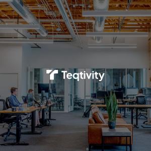 A modern, open-plan office space with exposed industrial ceiling. The Teqtivity logo is prominently displayed on a white wall. The space features standing desks with computer monitors, ergonomic office chairs, and employees working at their stations. A le