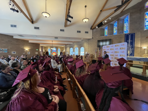 Side and back profile of three rows of graduates aged 18 - 60 years old sitting in a church in mahogany cap and gown.