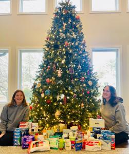 Becca and Laurie are two white women in their 40s. They are in their branded grey hoodies, sitting at the base of a christmas tree with basic essentials in a bin.