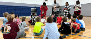 Children sitting on a gym floor listening to adults standing with holiday decorations.