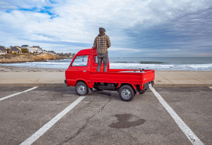 Subaru Sambar Truck parked by the ocean with surfer checking waves, showcasing the versatility of Japanese Kei trucks for beach and surf adventures.