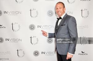 Photography of a man pointing to a logo on a backdrop. He is white, bald, and wears a grey suit with black buttergly tie. 