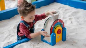 A young boy with spiky hair plays in a sandbox, pouring sand into a colorful interactive sand toy at Sandmagination