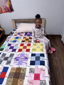 A little girl sits on a wooden twin size bed covered with a patchwork quilt.