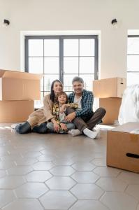 Family sits on the floor of a brand new home. They are surrounded by boxes and are sitting in front of big beautiful windows.