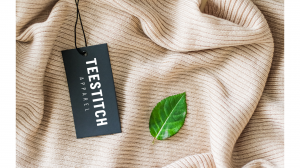 A close-up of a black TeeStitch Apparel clothing tag resting on a beige textured fabric, accompanied by a green leaf, symbolizing sustainability and eco-friendly fashion.